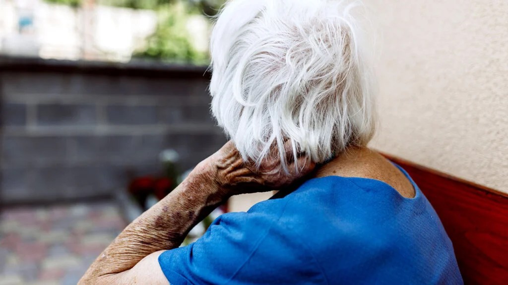 An older woman with gray hair wearing a blue top sits outside on a patio with her back facing the camera